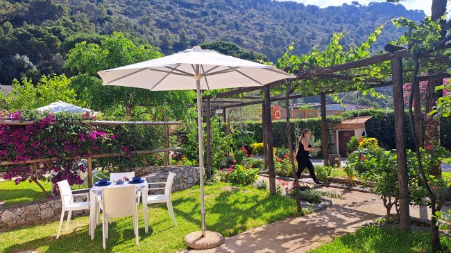 Giardino con tavolo, sedie bianche e ombrellone, circondato da fiori e colline verdi.