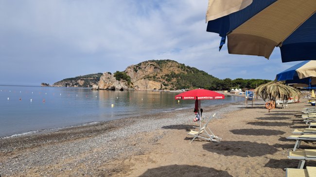 Spiaggia con sedie a sdraio, ombrelloni e mare calmo con scogli sullo sfondo.