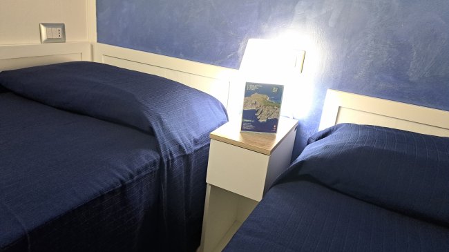 Due letti singoli con copriletto blu, separati da un comodino con una lampada accesa.