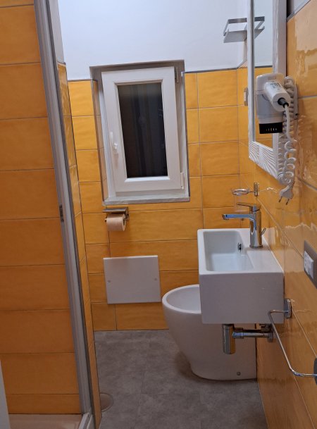 Bagno con piastrelle gialle, lavandino bianco, WC, finestra e doccia.