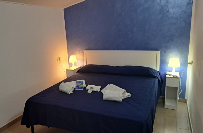 Camera da letto con parete blu, letto matrimoniale, due lampade e kit cortesia sul letto.