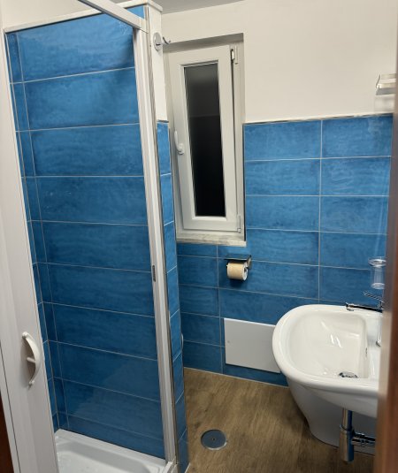 Bagno moderno con piastrelle blu, doccia, finestra stretta e lavandino bianco.