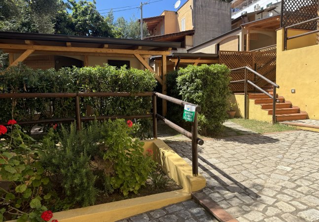 Ingresso con vialetto in pietra, aiuola con fiori rossi e una rampa di scale.
