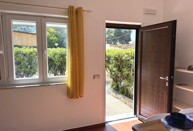 Porta aperta verso giardino, accanto a finestra con tende gialle.