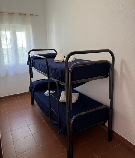 Stanza semplice con letto a castello e asciugamani su ciascun letto, finestra con tende leggere.