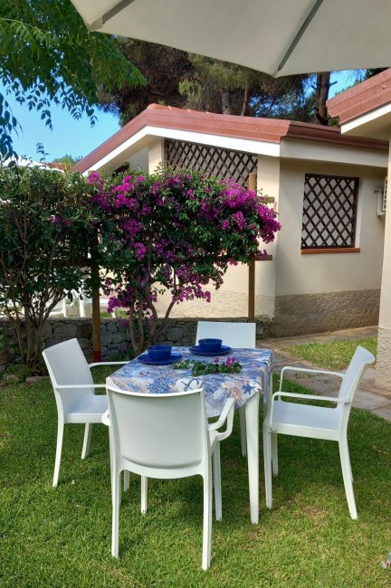 Giardino con tavolo e sedie bianche sotto un ombrellone, fiori viola decorano l'ambiente.