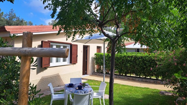Tavolo con sedie sotto un albero, vicino a una piccola casa beige con finestre e persiane marroni.