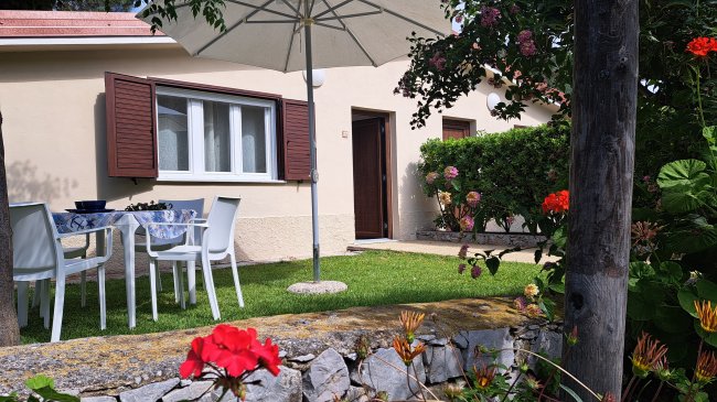Casa beige con tavolo e sedie bianche sotto un ombrellone, circondata da fiori colorati.