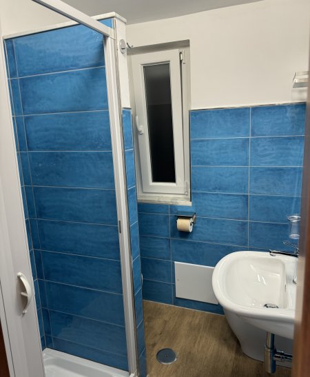 Bagno con piastrelle blu, doccia, finestra, lavandino e porta carta igienica.