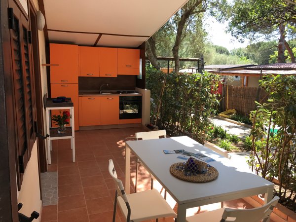 Una veranda con cucina arancione, tavolo bianco e sedie, circondata da piante.