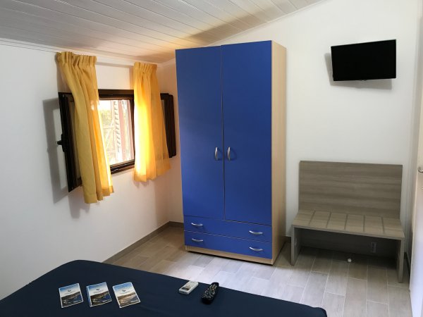 Camera con armadio blu, tenda gialla, TV a muro e brochure sul letto.