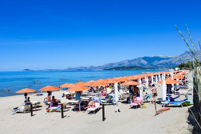 Spiaggia con file di ombrelloni arancioni, persone sulla sabbia, mare e montagne sullo sfondo.