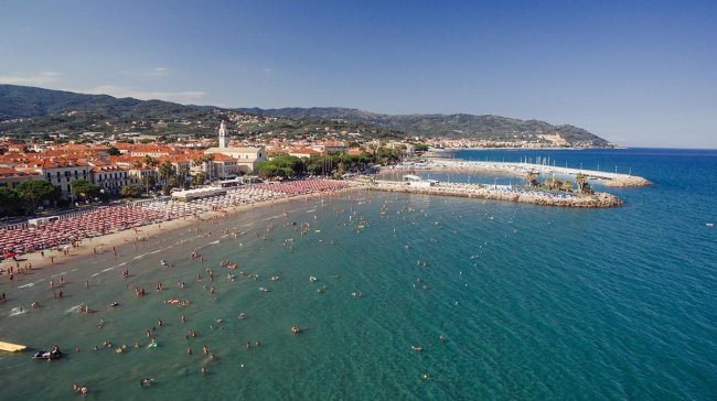 Spiaggia affollata con mare calmo e città sullo sfondo.