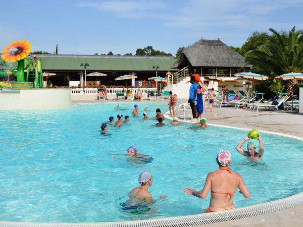 Persone in piscina giocano a palla, ombrelloni sullo sfondo.