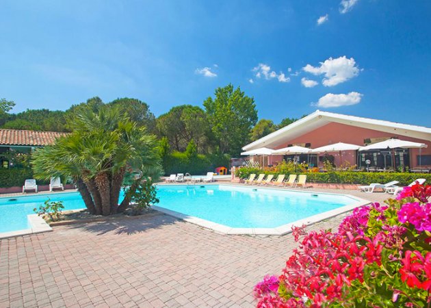 Piscina blu limpida con lettini e palme, un caffè rosa sullo sfondo in una giornata soleggiata.