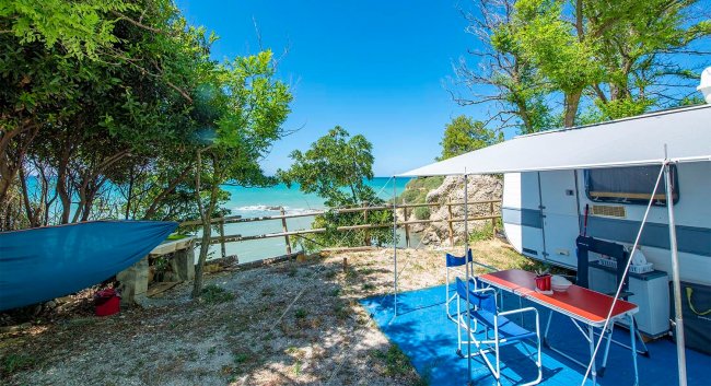 Camper con tavolo e vista mare tra alberi.