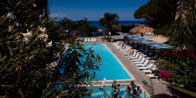 Piscina circondata da piante, vista mare, lettini bianchi.