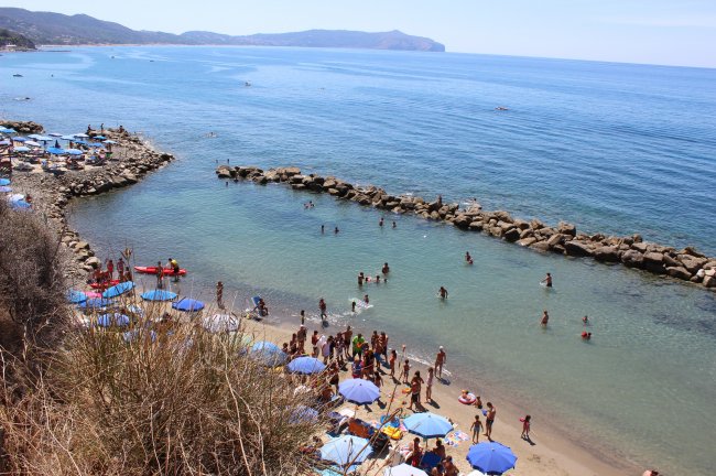 Spiaggia affollata con ombrelloni blu, persone in acqua e scogli sullo sfondo.