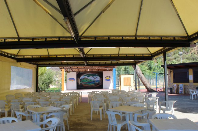 Uno spazio coperto con tavoli e sedie bianche, un palco con murale dipinto sullo sfondo.