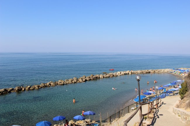 Spiaggia con ombrelloni blu, mare calmo e persone che nuotano; barriera di rocce in lontananza.