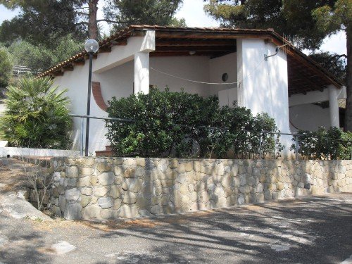 Casa bianca con tetto marrone, circondata da cespugli e alberi.