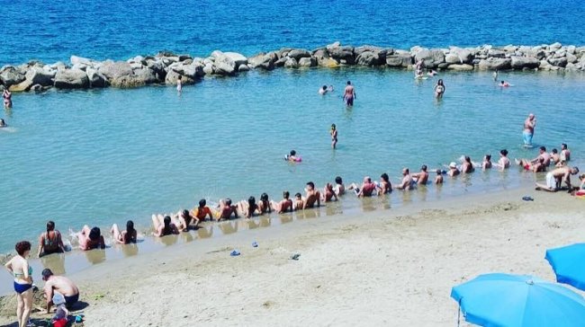 Persone sedute sulla sabbia e in acqua vicino a una barriera rocciosa in spiaggia.