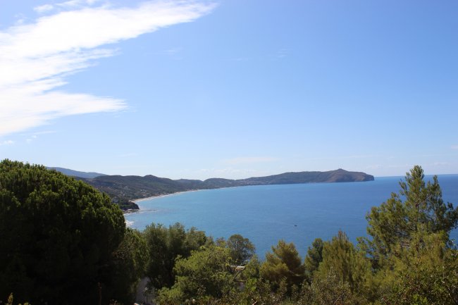 Una costa con un mare blu, circondata da verdi colline e alberi sotto un cielo sereno.
