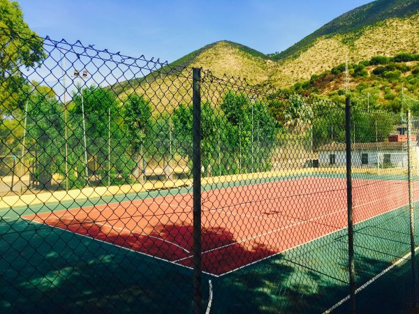 Un campo da tennis recintato, con montagne verdi sullo sfondo sotto un cielo azzurro.