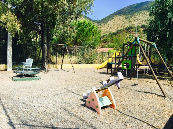 Area giochi all'aperto con scivolo, altalene e giostra su ghiaia, circondata da colline.