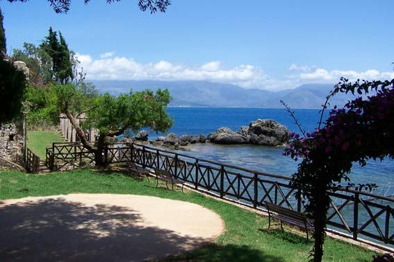 Vista su un mare blu con scogli, prato verde, panchine e pergola fiorita.