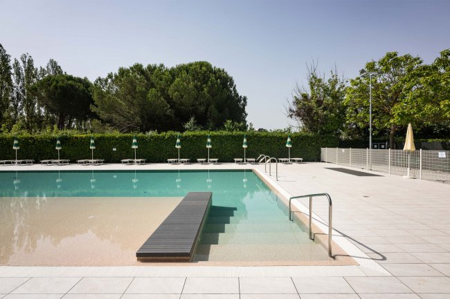 Piscina con passerella e ombrelloni verdi sullo sfondo.