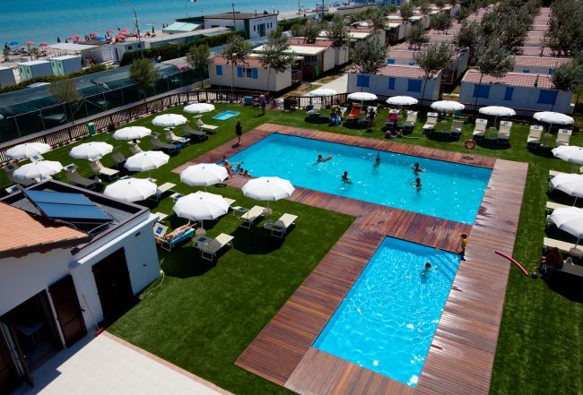 Piscina all'aperto con persone che nuotano, ombrelloni bianchi e sdraio, vicino al mare.