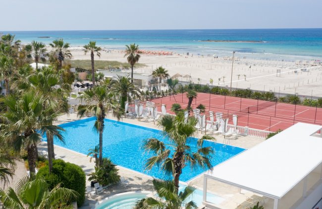 Vista su piscina, campo da tennis e spiaggia con mare azzurro sullo sfondo.