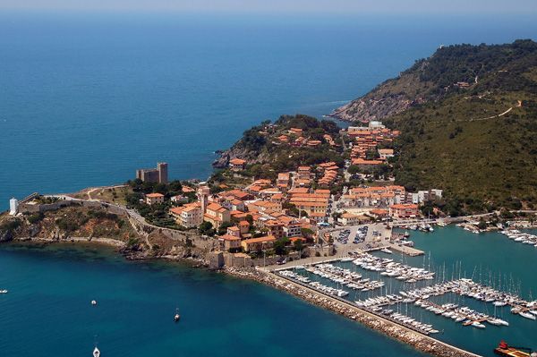 Vista aerea di un borgo costiero con porto, circondato dal mare e colline.