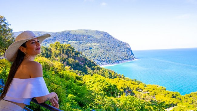 Donna con cappello ammira un paesaggio costiero verde e blu.