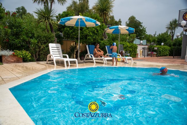 Piscina con sdraio e ombrelloni bianchi e blu, persone sedute e in acqua.