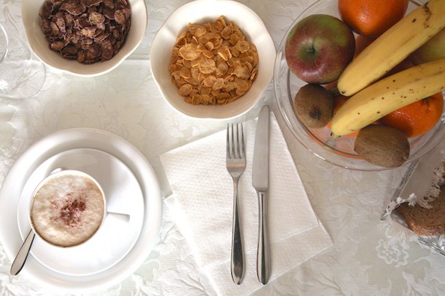 Colazione con cereali, cappuccino e frutta su una tovaglia bianca.