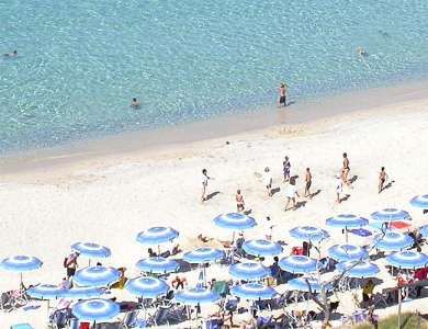 Spiaggia con lettini e ombrelloni blu disposti ordinatamente, persone che camminano lungo la riva.