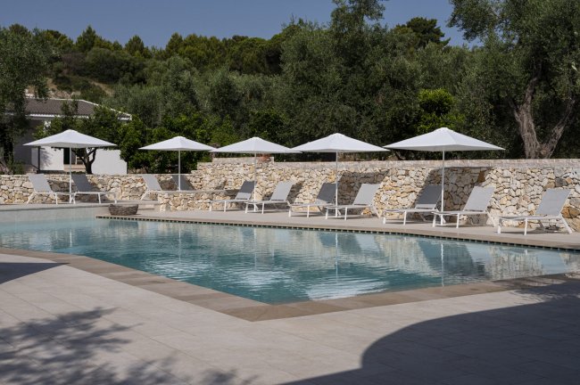 Piscina con lettini bianchi e ombrelloni, circondata da un muro di pietra.