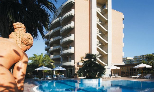 Edificio moderno con balconi curvi, piscina circondata da sedie a sdraio, ombrelloni e palme.