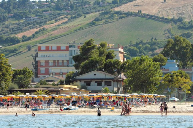 Spiaggia affollata con colline e edifici sullo sfondo.