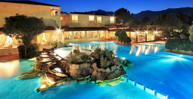 Piscina illuminata con cascate e vegetazione, edificio sullo sfondo.