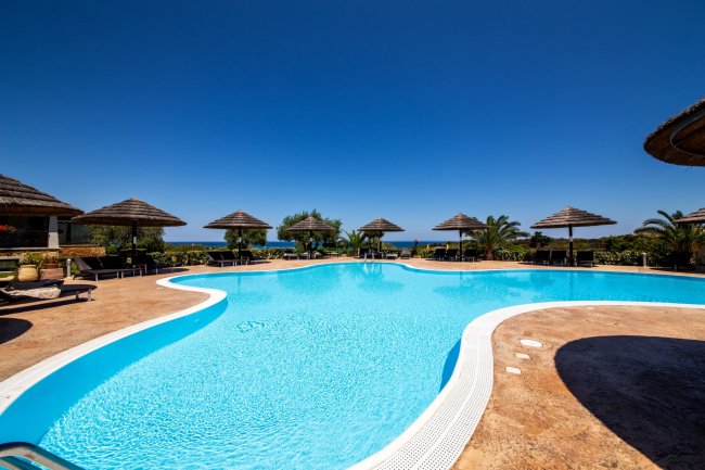 Piscina all'aperto con ombrelloni di paglia e cielo azzurro.