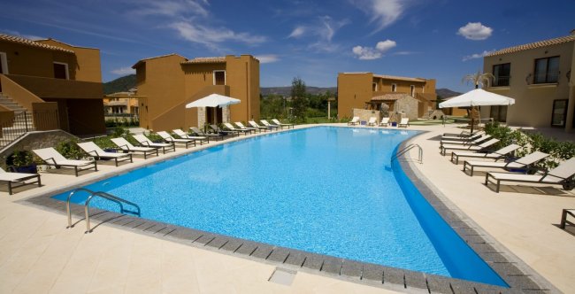 Piscina con lettini, ombrelloni e edifici sullo sfondo.
