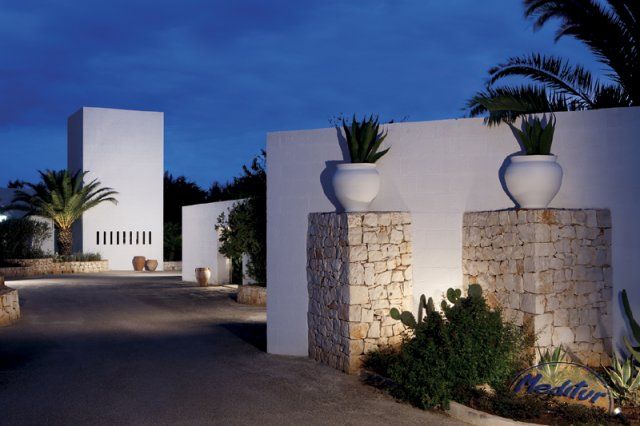 Ingresso esterno con muri bianchi, vasi grandi, palme e cielo blu scuro.