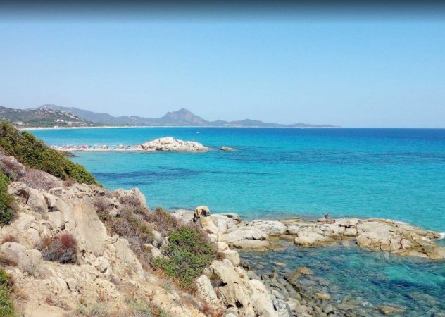 Costa rocciosa con mare turchese e colline lontane.