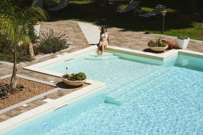 Piscina con donna su gradino immersa, circondata da piante e lettini.
