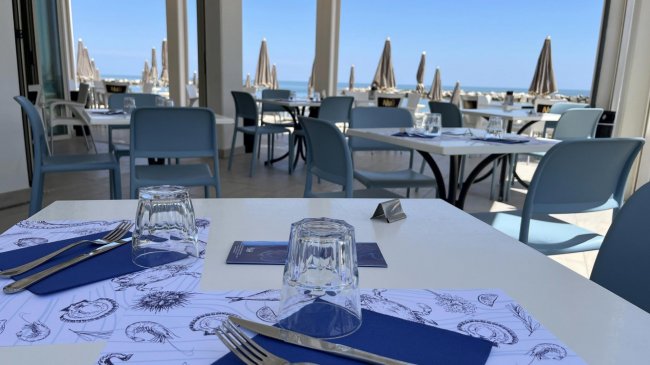 Tavoli apparecchiati in un ristorante all'aperto con vista mare e sedie blu.