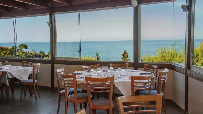 Sala da pranzo con tavoli apparecchiati, sedie in legno e grandi finestre con vista mare.