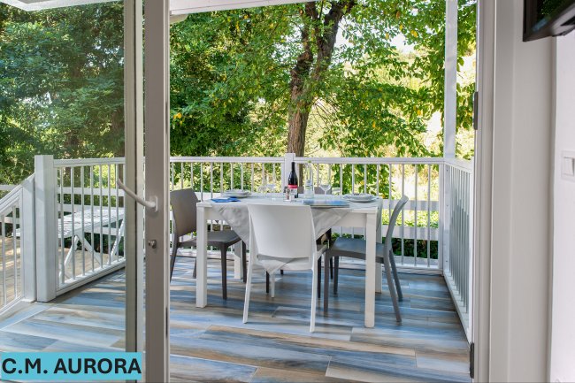 Porta aperta su una veranda con tavolo apparecchiato e ringhiera bianca immersa nel verde.
